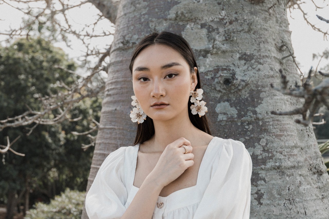 a woman standing in front of a tree wearing white floral earrings