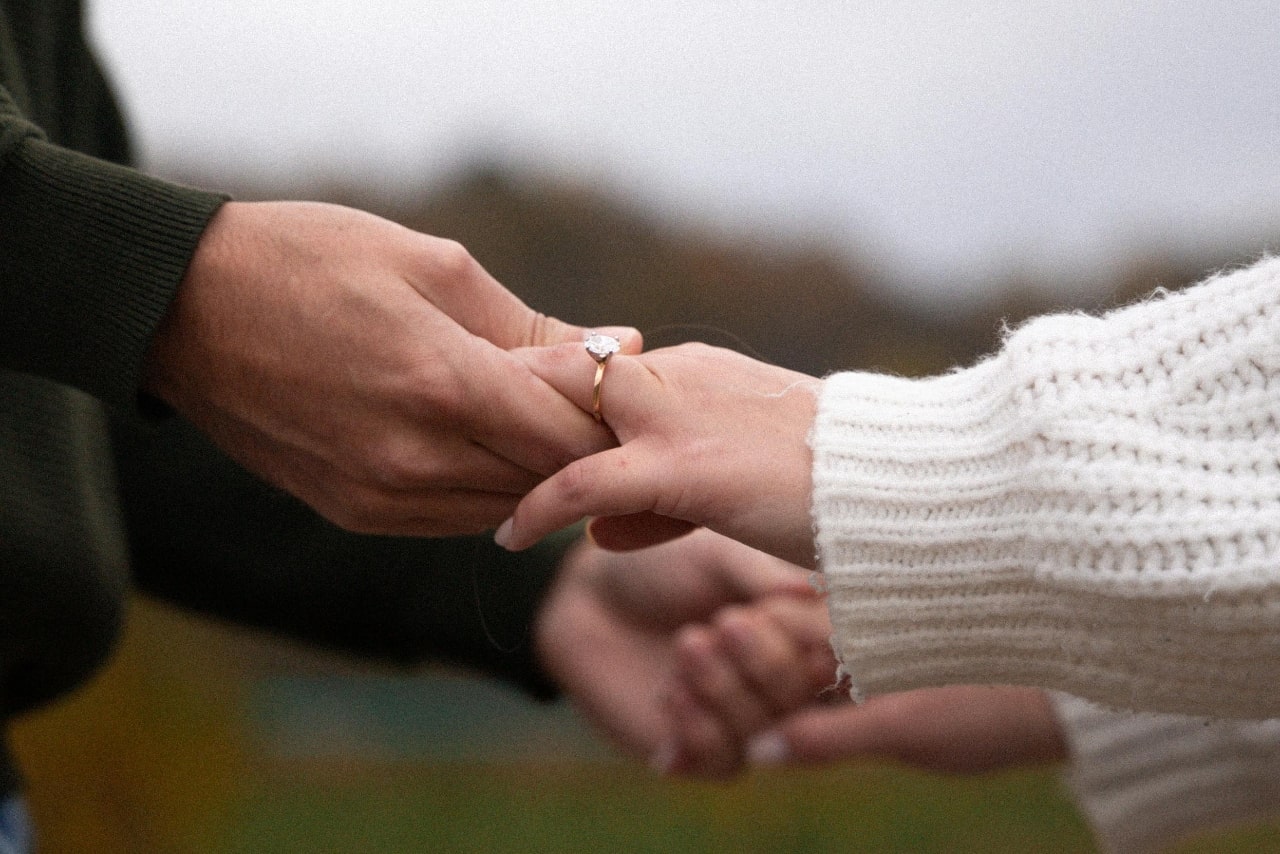 A close up of two intertwined hands, the woman wearing a white knitted sweater and the man in a khaki sweatshirt, showcasing yellow gold solitaire engagement rings.