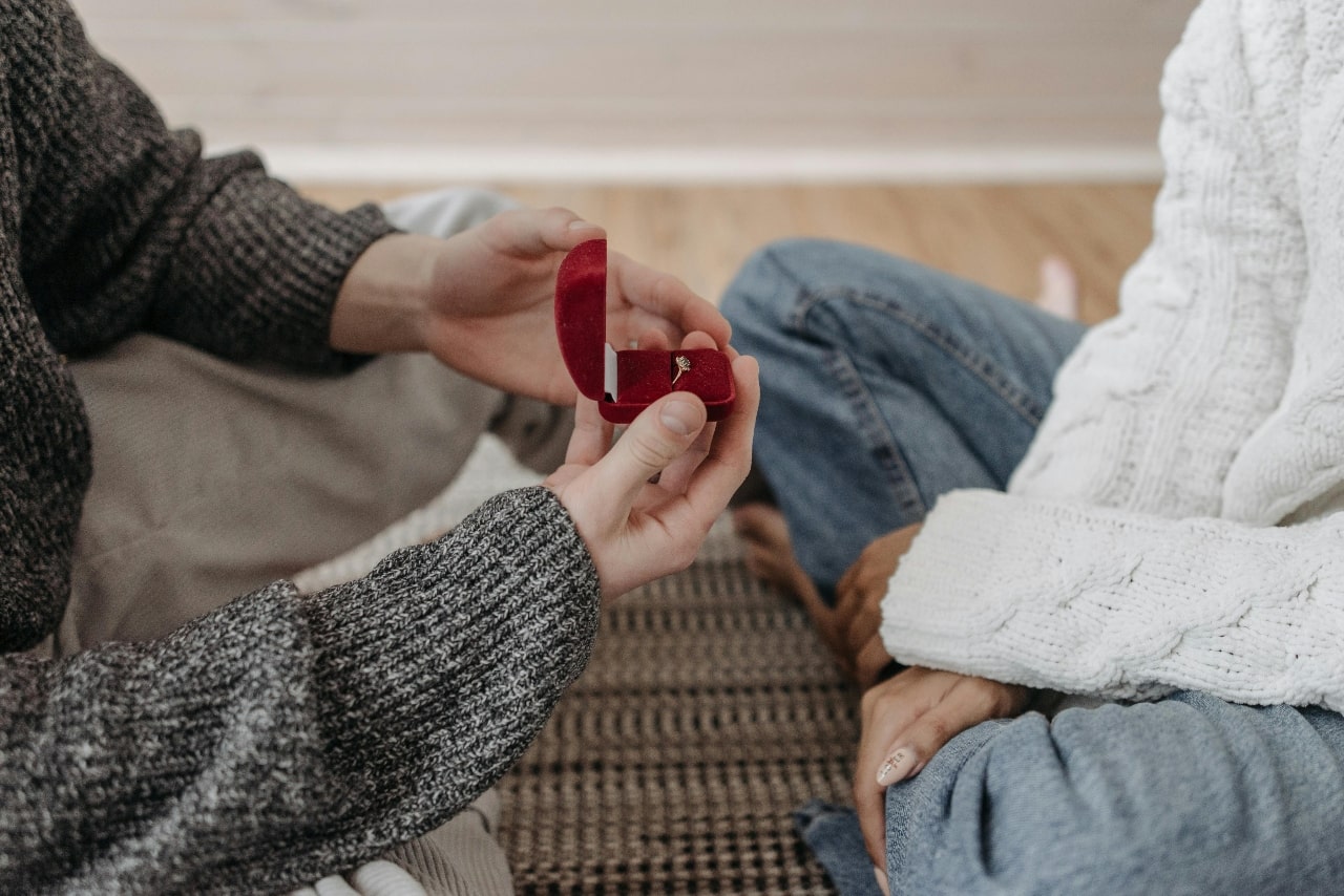 Two people sitting on the floor, with one proposing to another with an engagement ring in a red velvet box.