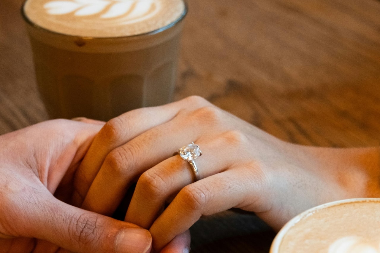 A close-up of a man holding a woman&rsquo;s hand as they drink lattes, with emphasis on the oval cut solitaire ring on her finger.