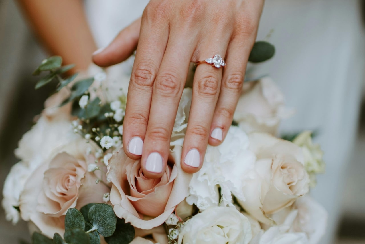 A three stone diamond engagement ring a woman’s hand on top of a bouquet of white roses.