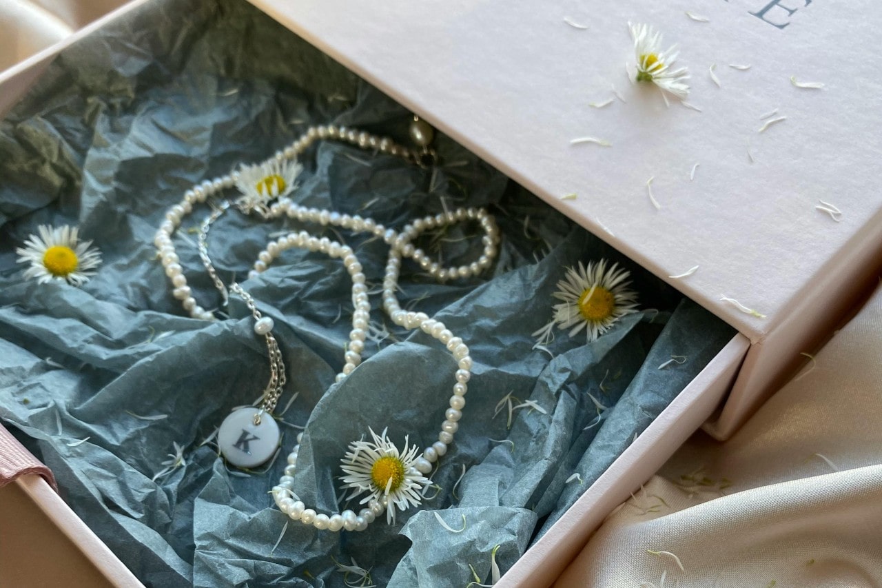 A delicate pearl and initial pendant necklace displayed in a gift box filled with tissue paper and flowers.