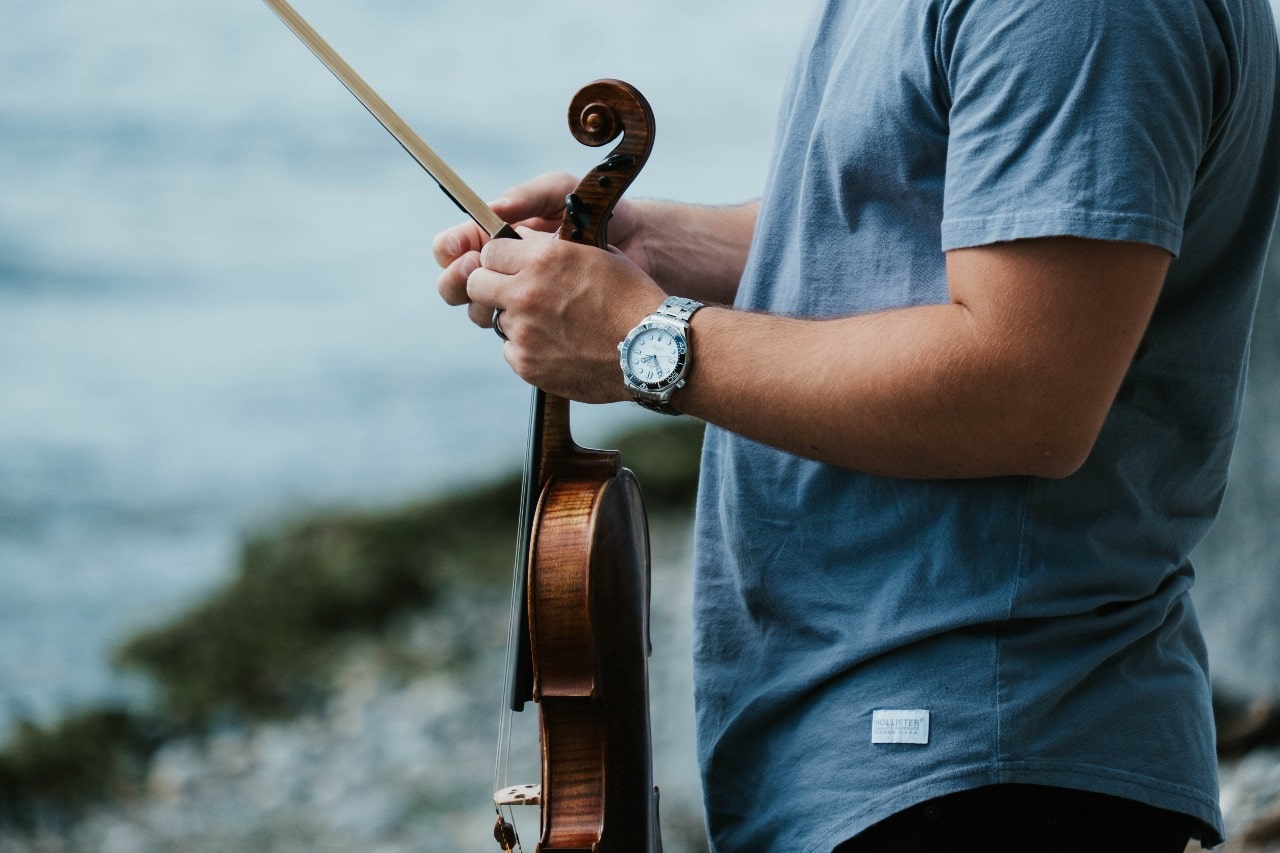 A close-up of a young man holding a violin and wearing a stainless steel sports watch.