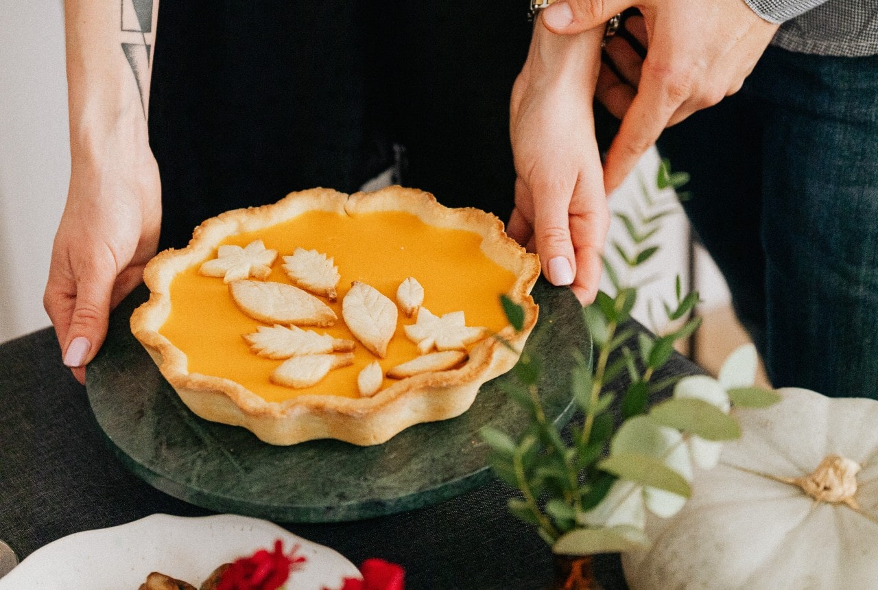 A woman sets down a decorative pie on a Thanksgiving table while a man supports her wrist.