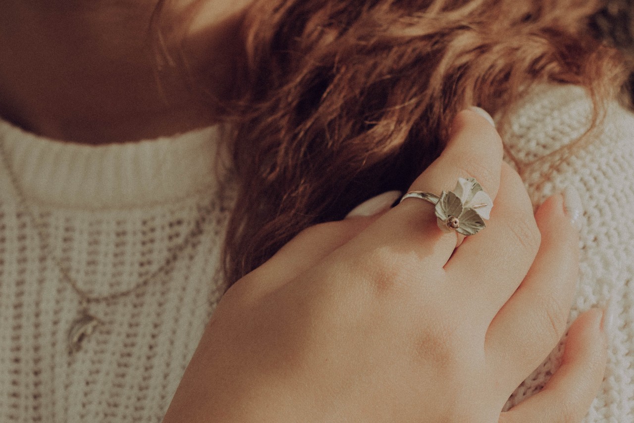 A woman in a sweater touches her hair, revealing her fashion ring and necklace.