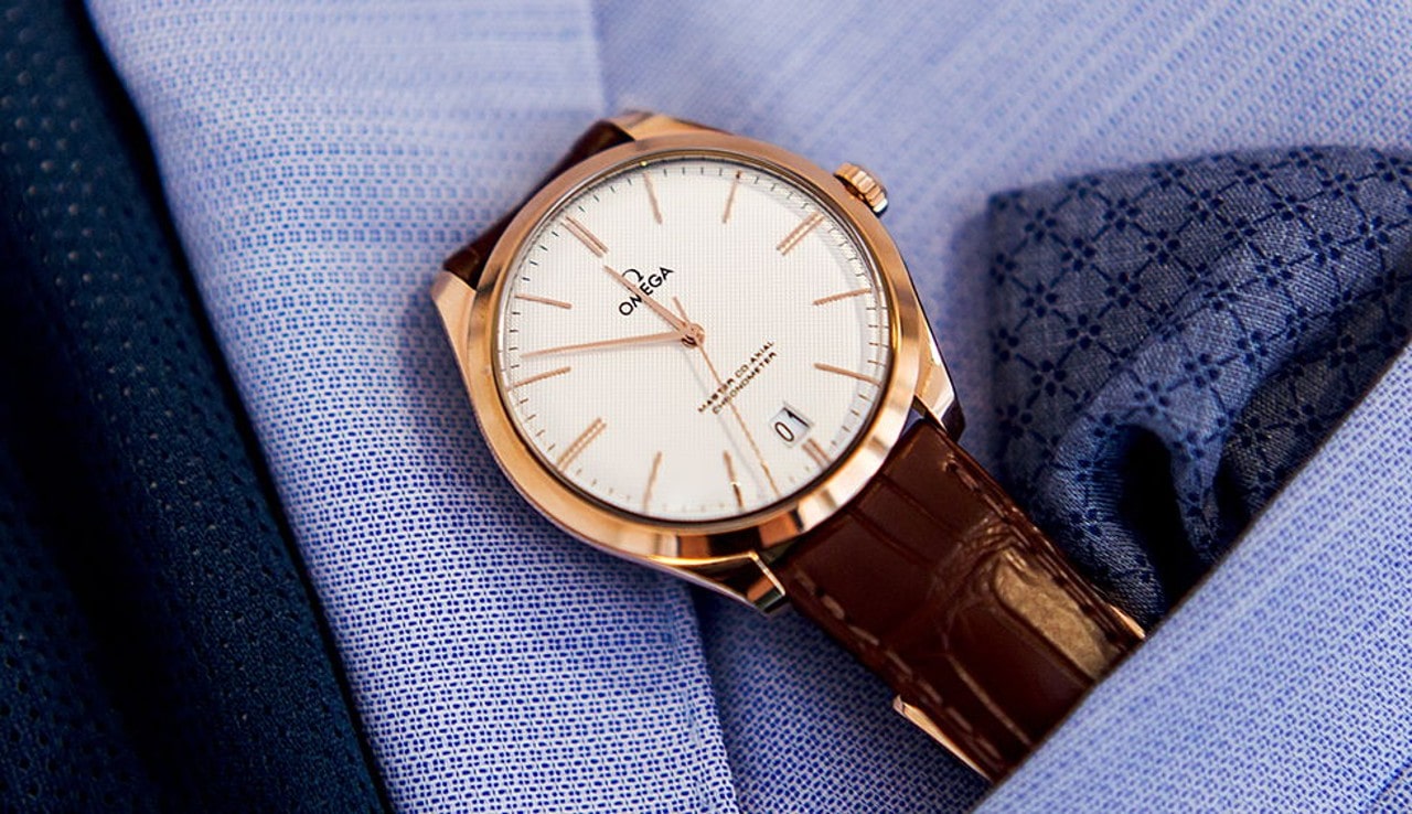 An Omega wristwatch with a brown leather strap and gold case, resting on a blue suit and pocket square.