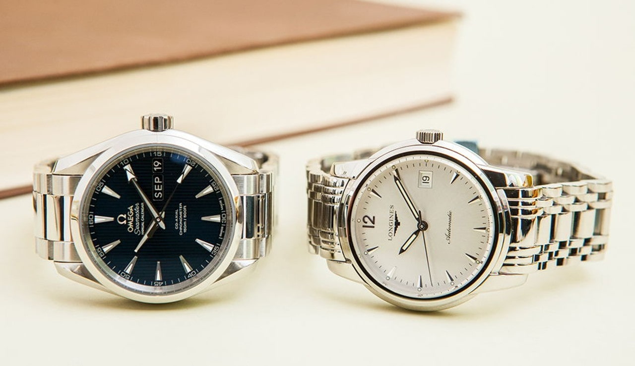 Two elegant wristwatches, an Omega with a dark dial and a Longines with a white dial, placed side by side on a light surface.