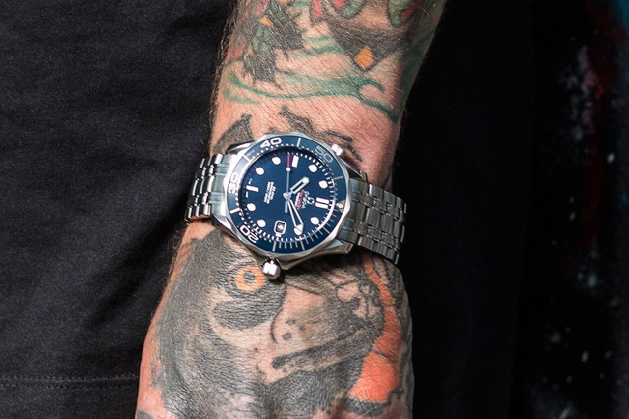 A tattooed wrist adorned with a stainless steel dive watch featuring a deep blue dial, luminous markers, and a rotating bezel, set against a dark background.