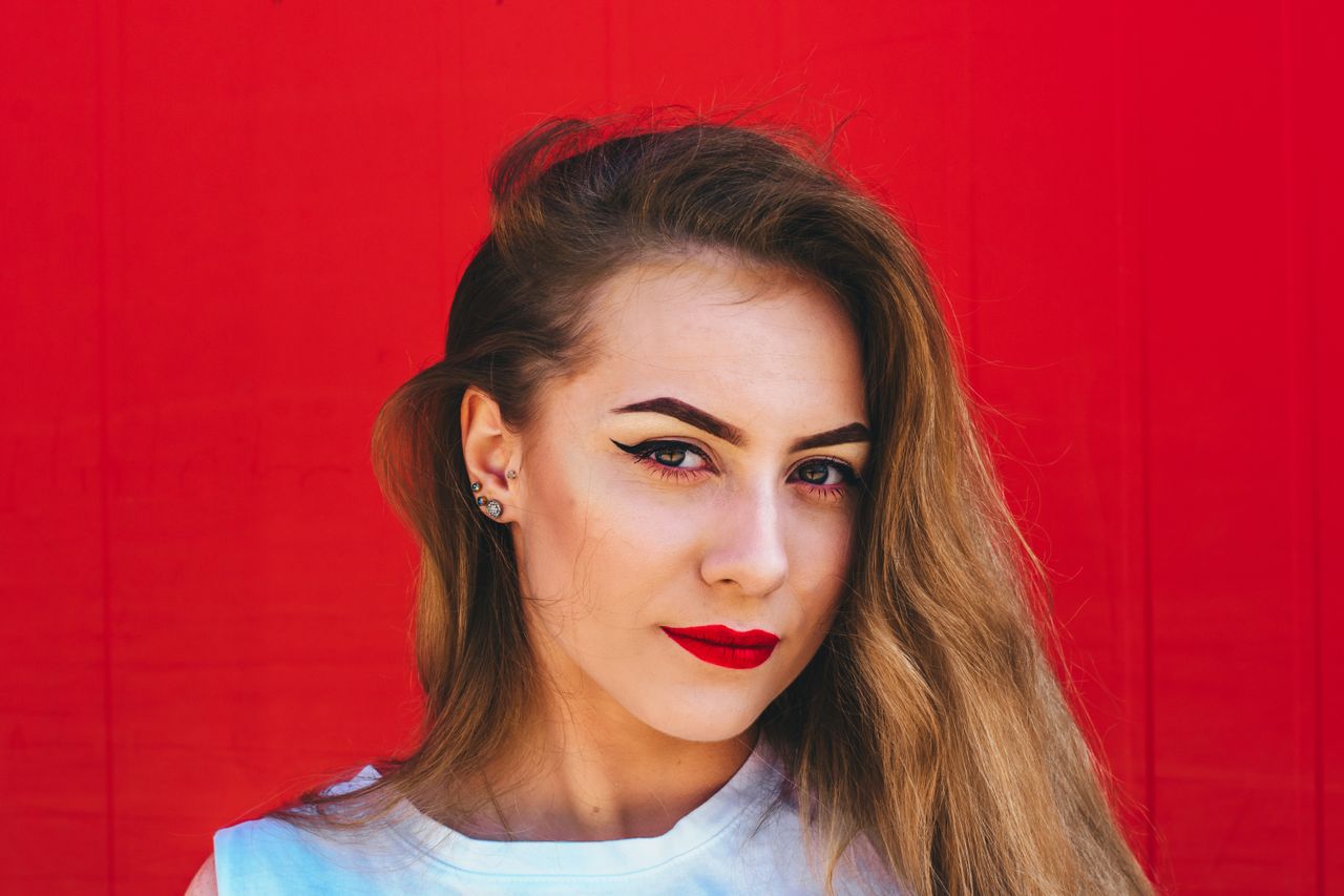 A woman wearing red lipstick sports multiple stud earrings against a red wall.
