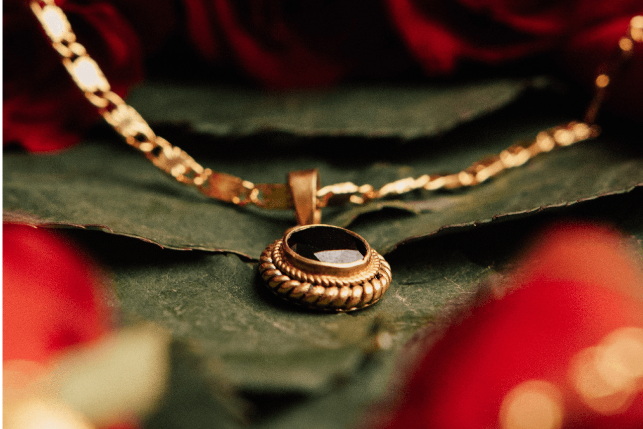 A gold necklace with a dark gemstone pendant lies on a green leaf, surrounded by soft-focus red roses.