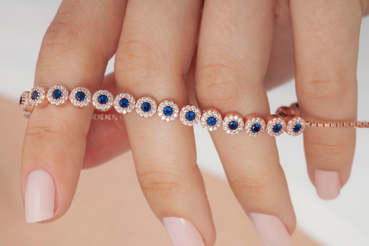 A hand with pale pink manicured nails holds a rose gold bracelet adorned with round blue sapphires.