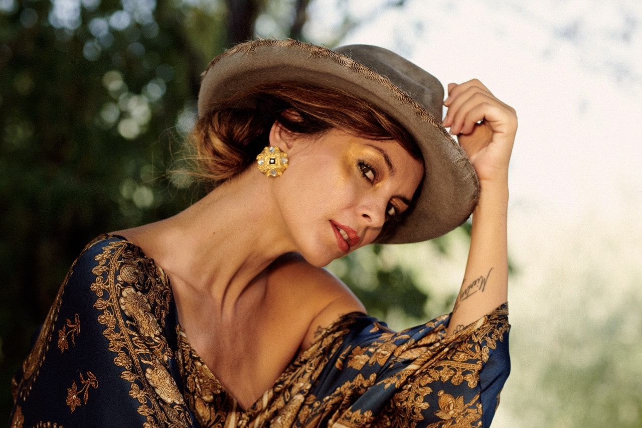 A woman wearing a wide-brimmed hat and a patterned off-shoulder top with bold gold earrings poses outdoors.