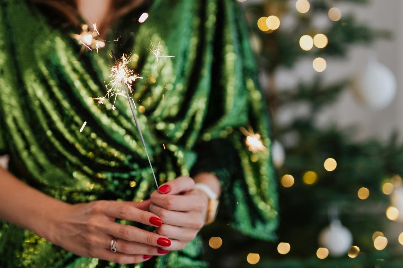 A person in a glittery green dress wearing rings and watch, holds a lit sparkler with red-painted nails, standing in front of a softly lit Christmas tree.