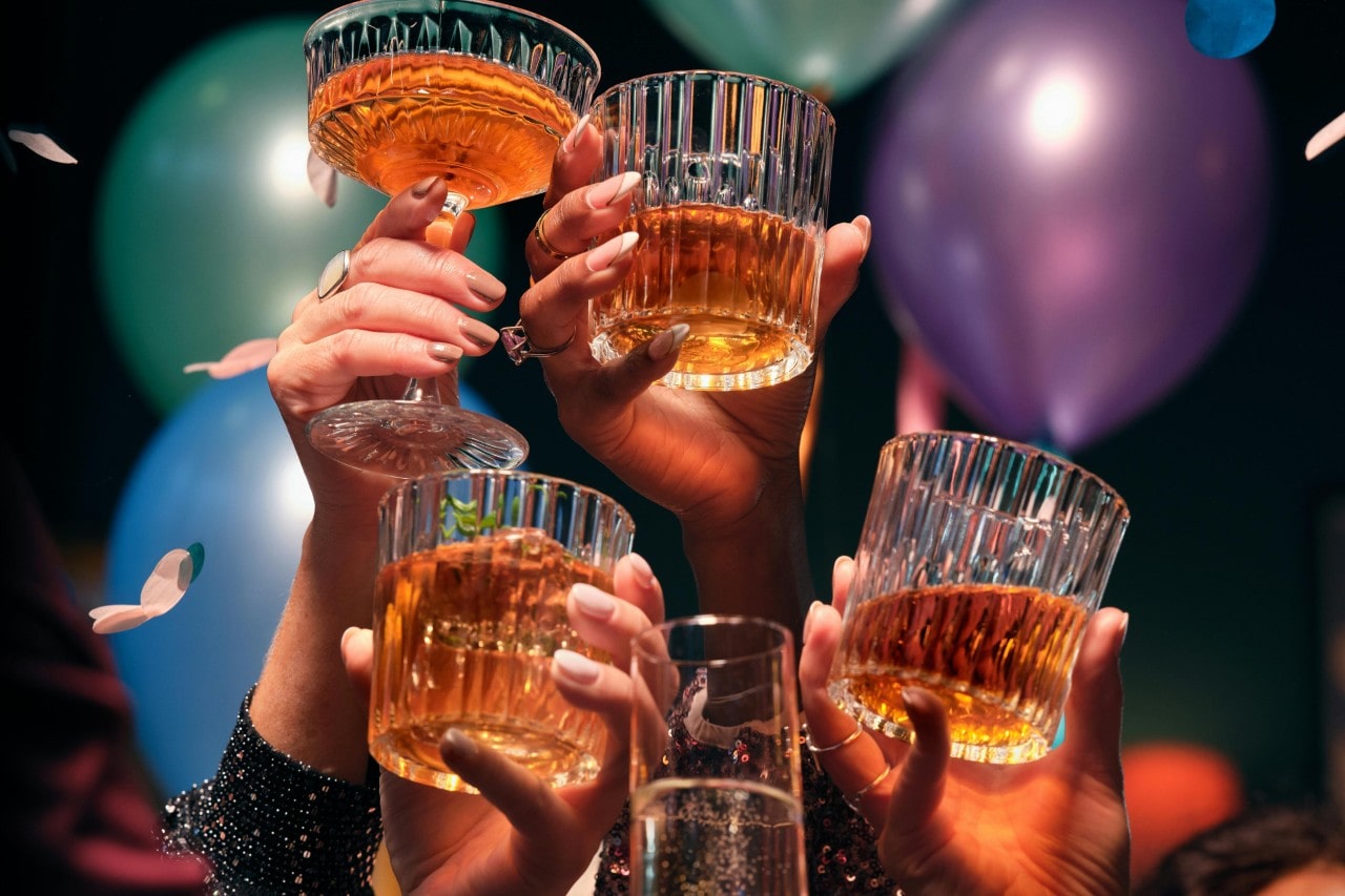 People’s hands raising a toast at a New Year’s Eve party, surrounded by balloons and confetti.