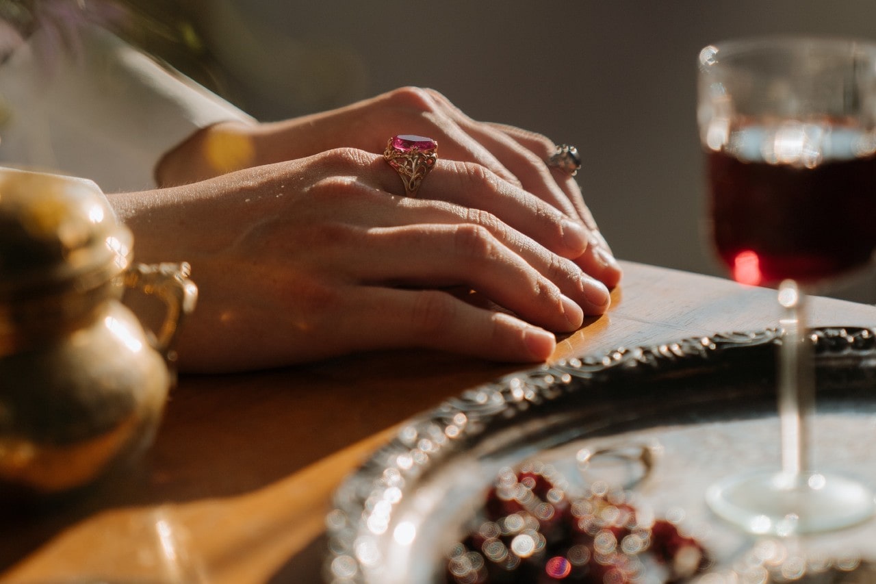 a pair of hand sitting on a table, wearing gemstone rings next to a glass of wine