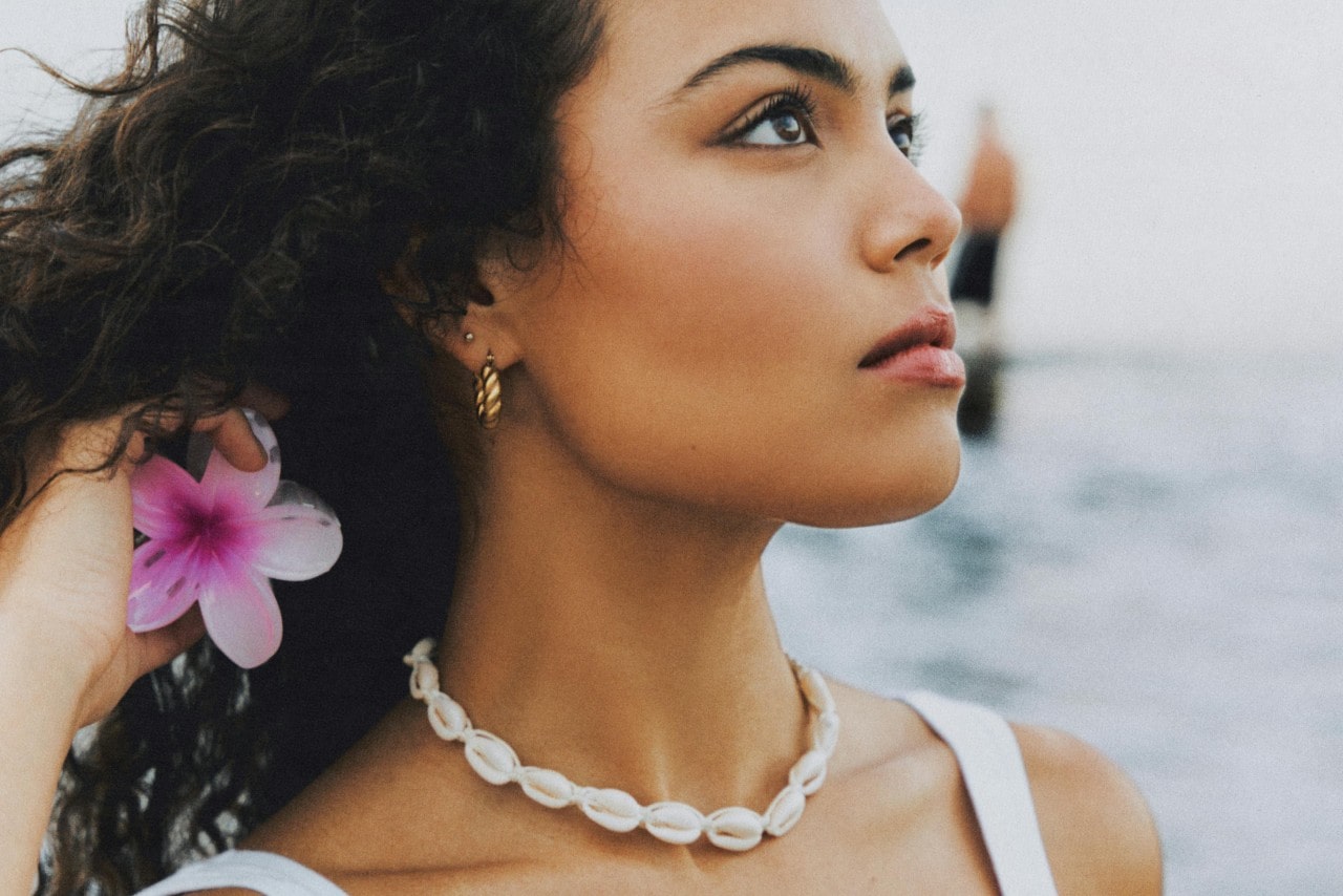A woman on the beach wearing a shell necklace and holding a flower in her hair.