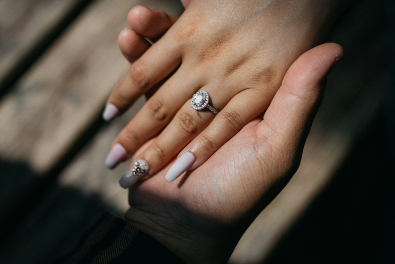 a man’s hand holding a woman’s in the sunlight, wearing a halo engagement ring