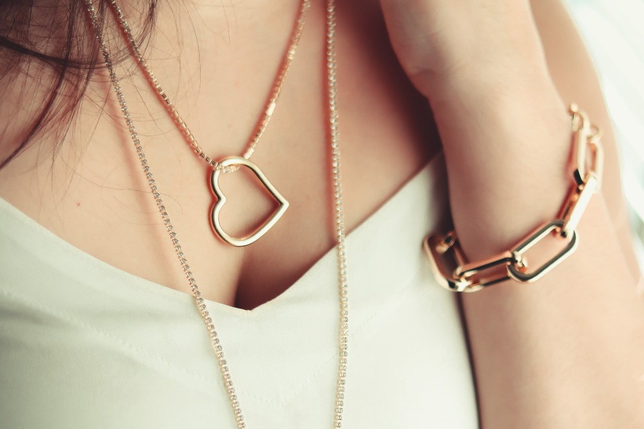 Close-up of a woman's chest in a white top showcasing layered yellow gold heart necklaces, and a gold link bracelet.