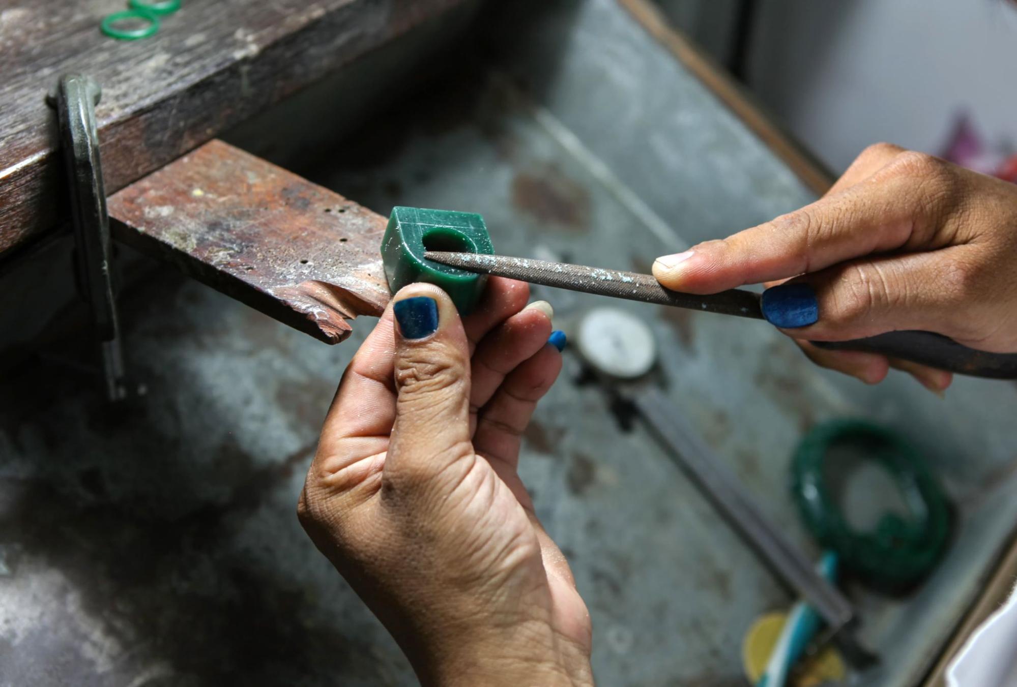 Hands with blue nail polish delicately filing a green wax ring on a jeweler's workbench.
