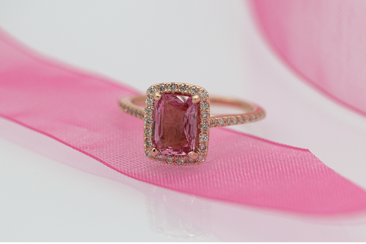 A close up of rose gold halo ring with pink center gemstone, accented with small diamonds along the band, on top of the pink ribbon.
