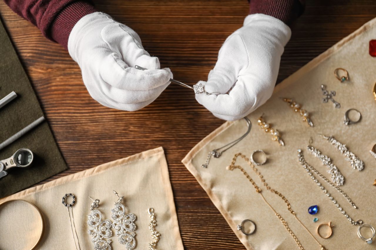 A person wearing white gloves examines a diamond ring with a magnifying tool on a wooden table, while nearby are trays displaying various pieces of jewelry.