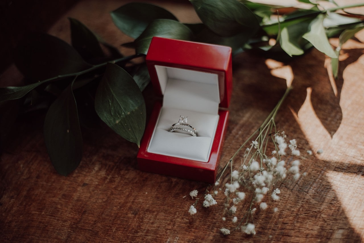A diamond solitaire white gold ring with a matching dainty eternity wedding band in a red ring box on the table surrounded by flowers.