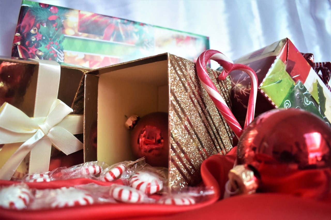 A close-up of a golden gift box tipped open on its side, with a single red ornament inside and peppermints strewn around the top.