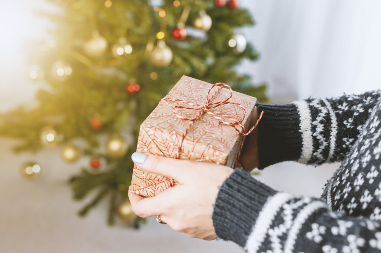 A close-up of a woman in a festive sweater holding an elegantly wrapped gift, a Christmas tree in the background.