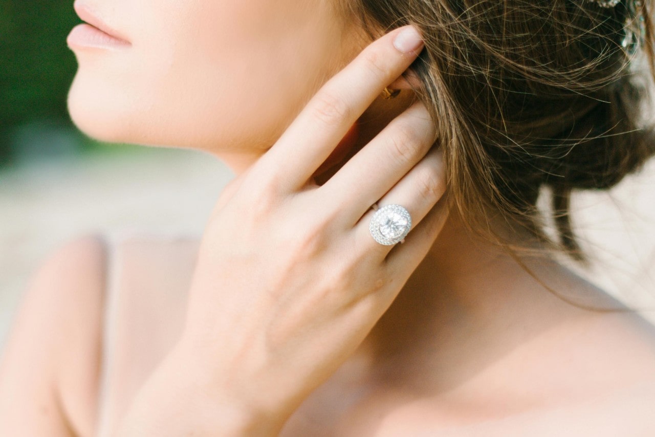 A close-up of a bride with her hand in her hair, wearing a large diamond halo engagement ring.