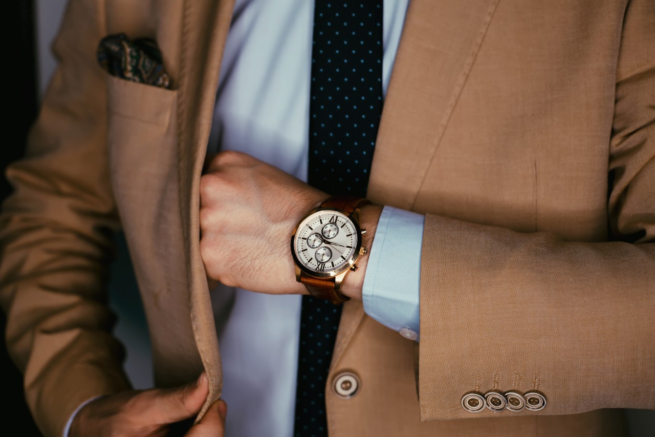 A man reached into the inside pocket of his beige suit jacket with a focus on his luxury watch with a GMT complication, yellow gold watch case, and leather strap.