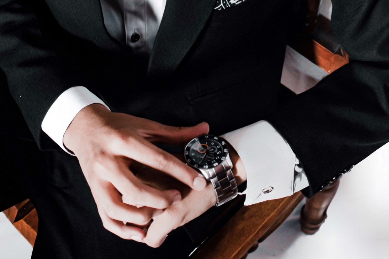 A man in a black suit showcasing a silver watch.