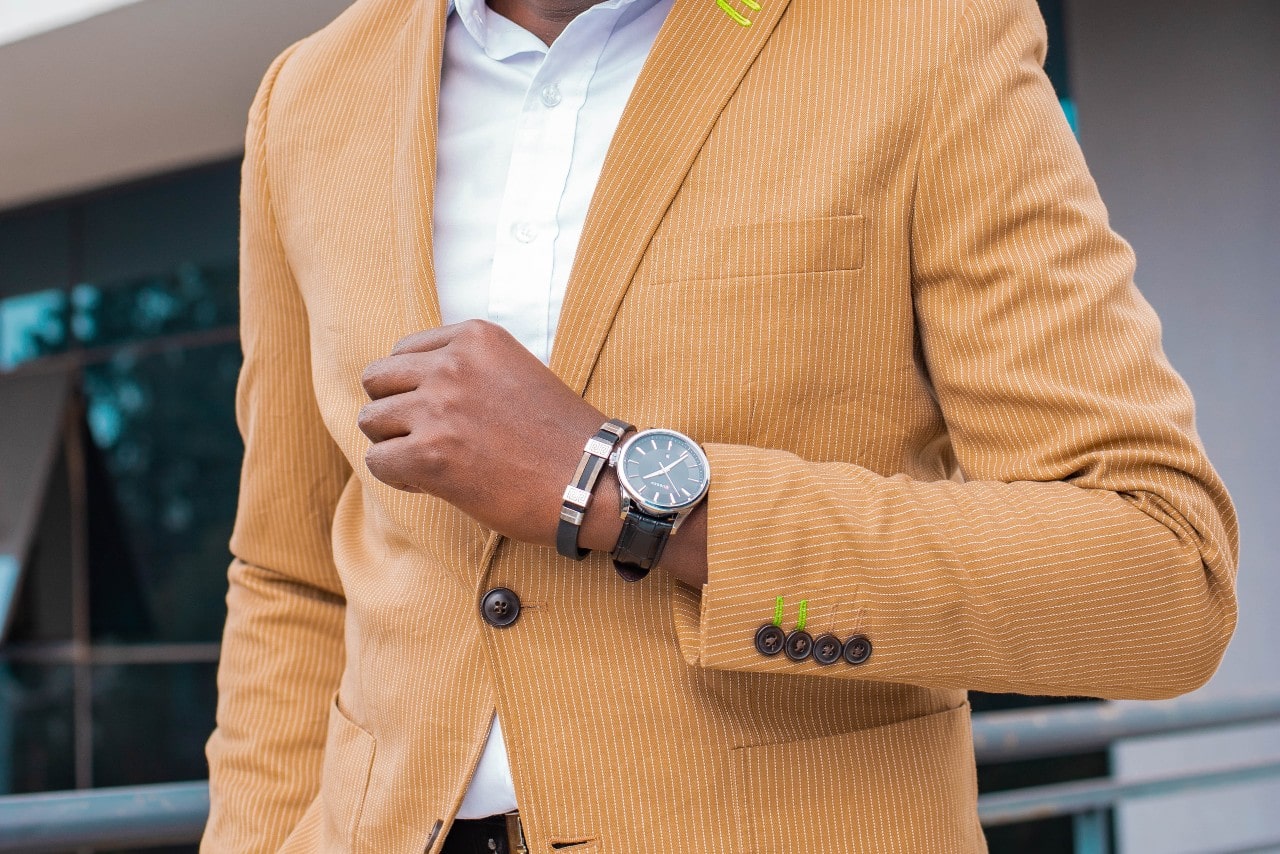A man in a mustard suite showcasing a black watch with a brown band.
