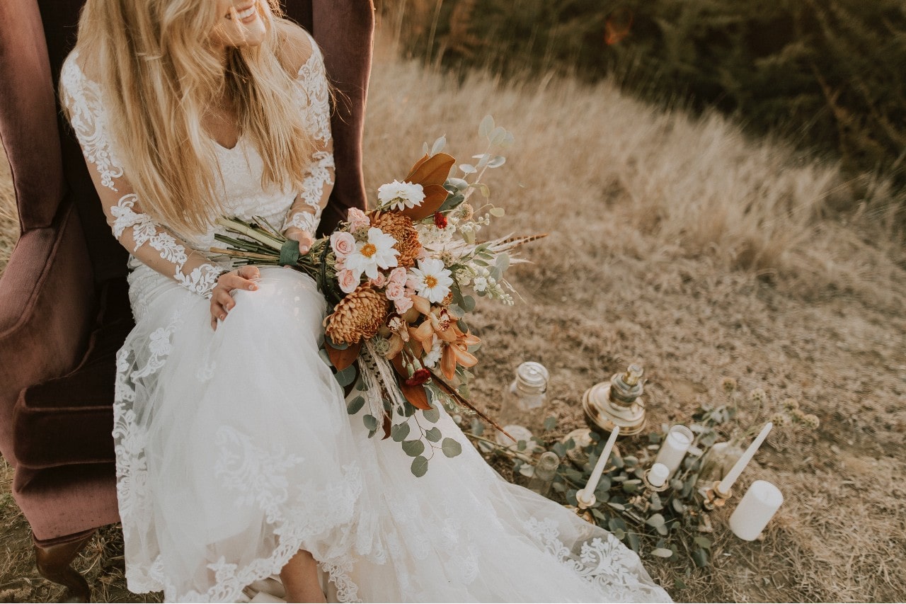 a bride sitting in a velvet chair outdoors, holding a bouquet of flowers