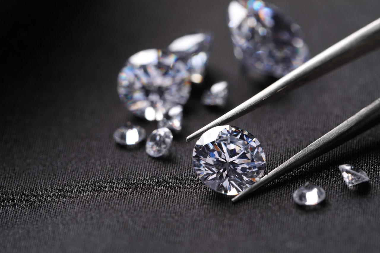 A close-up of multiple round cut diamonds of varying size, one emphasized with jeweler&rsquo;s tweezers, on a dark denim backdrop.