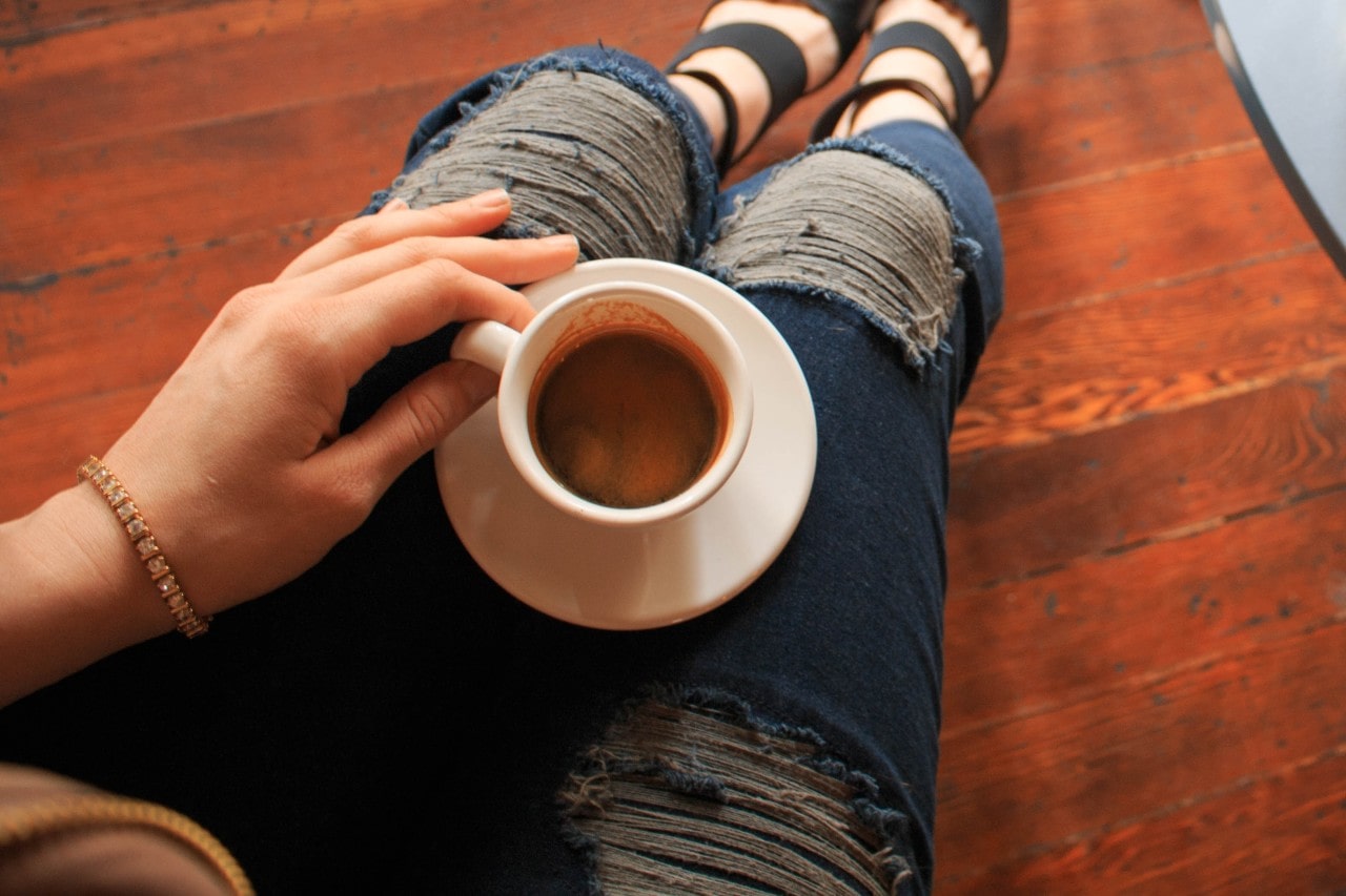 A woman in ripped jeans sitting on the floor sips coffee.