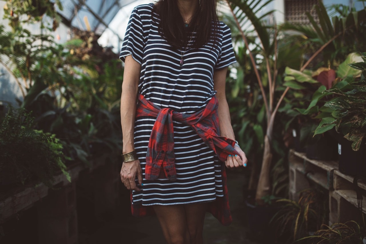 A woman in a t-shirt dress wearing jewelry walks through a greenhouse.