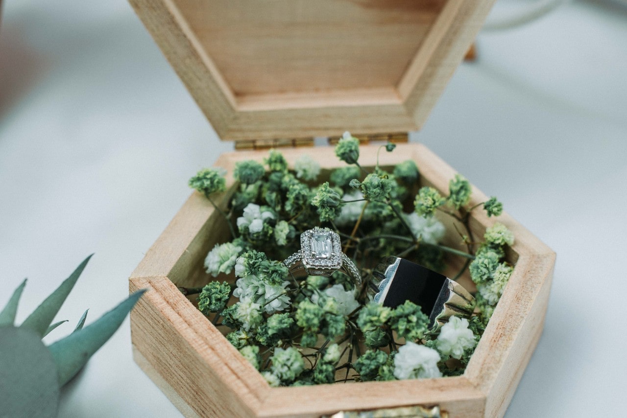 An emerald halo engagement ring sits in a variety of small white flowers in a wooden ring box.
