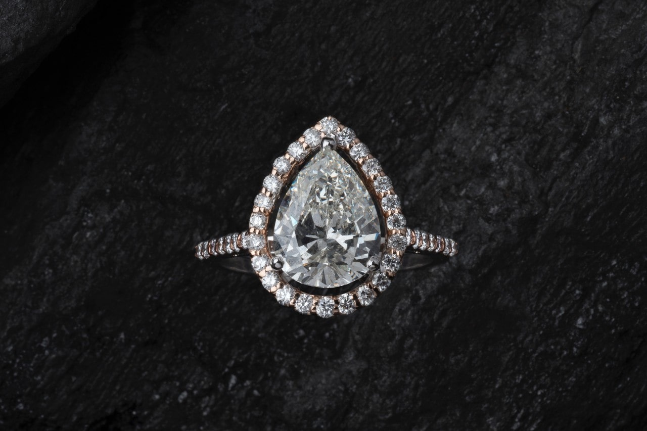 A platinum teardrop diamond halo engagement ring sits on a black surface. A platinum teardrop diamond halo engagement ring sits on a black surface.