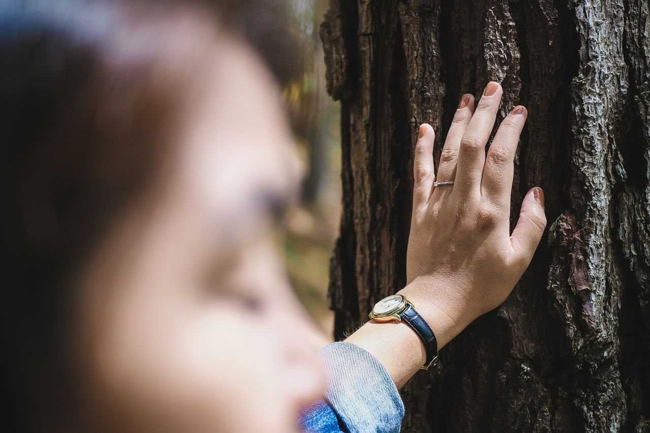 a woman leaning against a tree on a hike, showing off her side stone engagement ring.