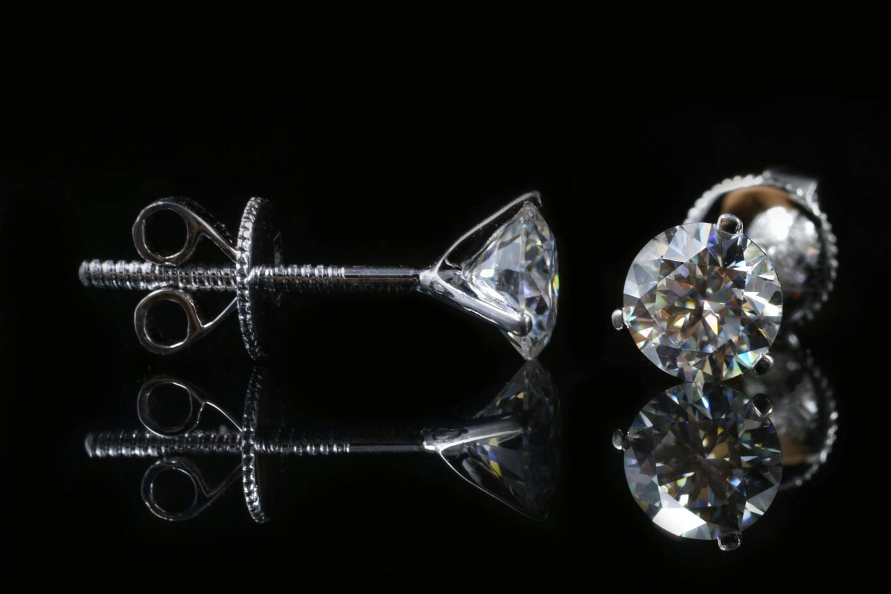 A close-up of a pair of diamond stud earrings on a black backdrop, one displayed from the front and the other from the side.