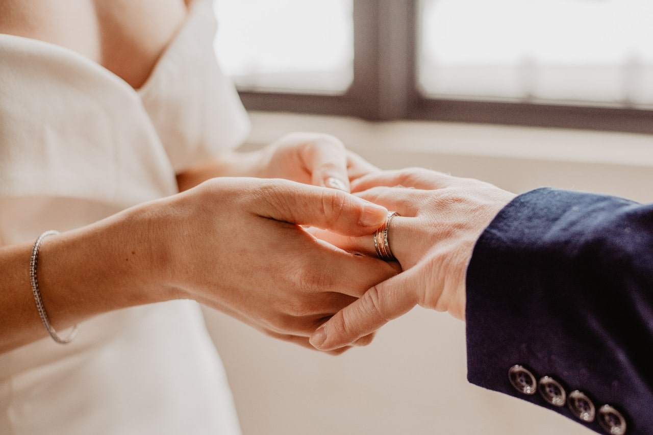 Close-up of a bride in a white dress placing a ring on a groom's finger, evoking a sense of warmth and commitment. Soft lighting enhances the tender moment.