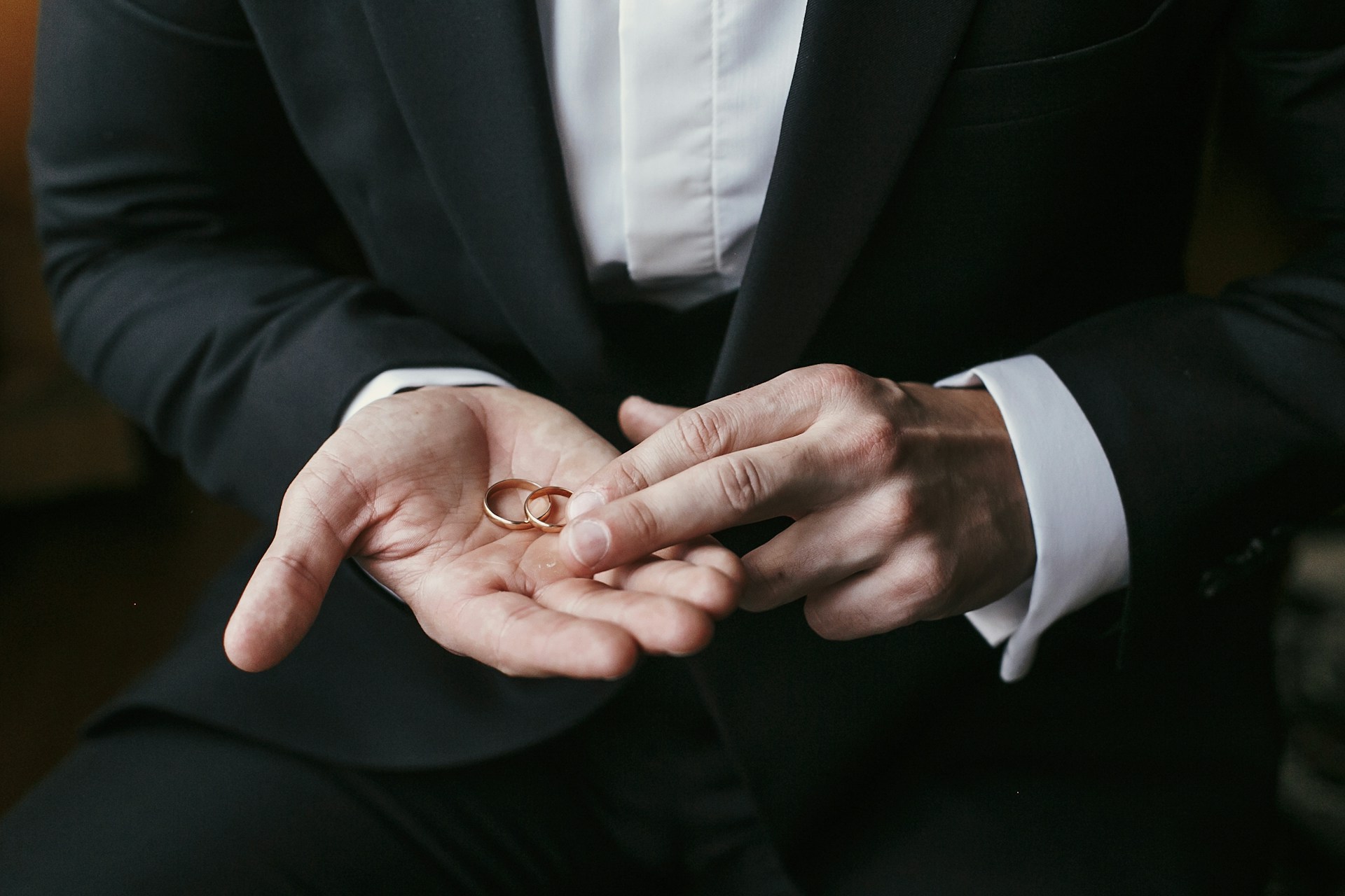 A man in a black suit gently holds two gold wedding rings in his open palm, conveying a sense of anticipation and solemnity.