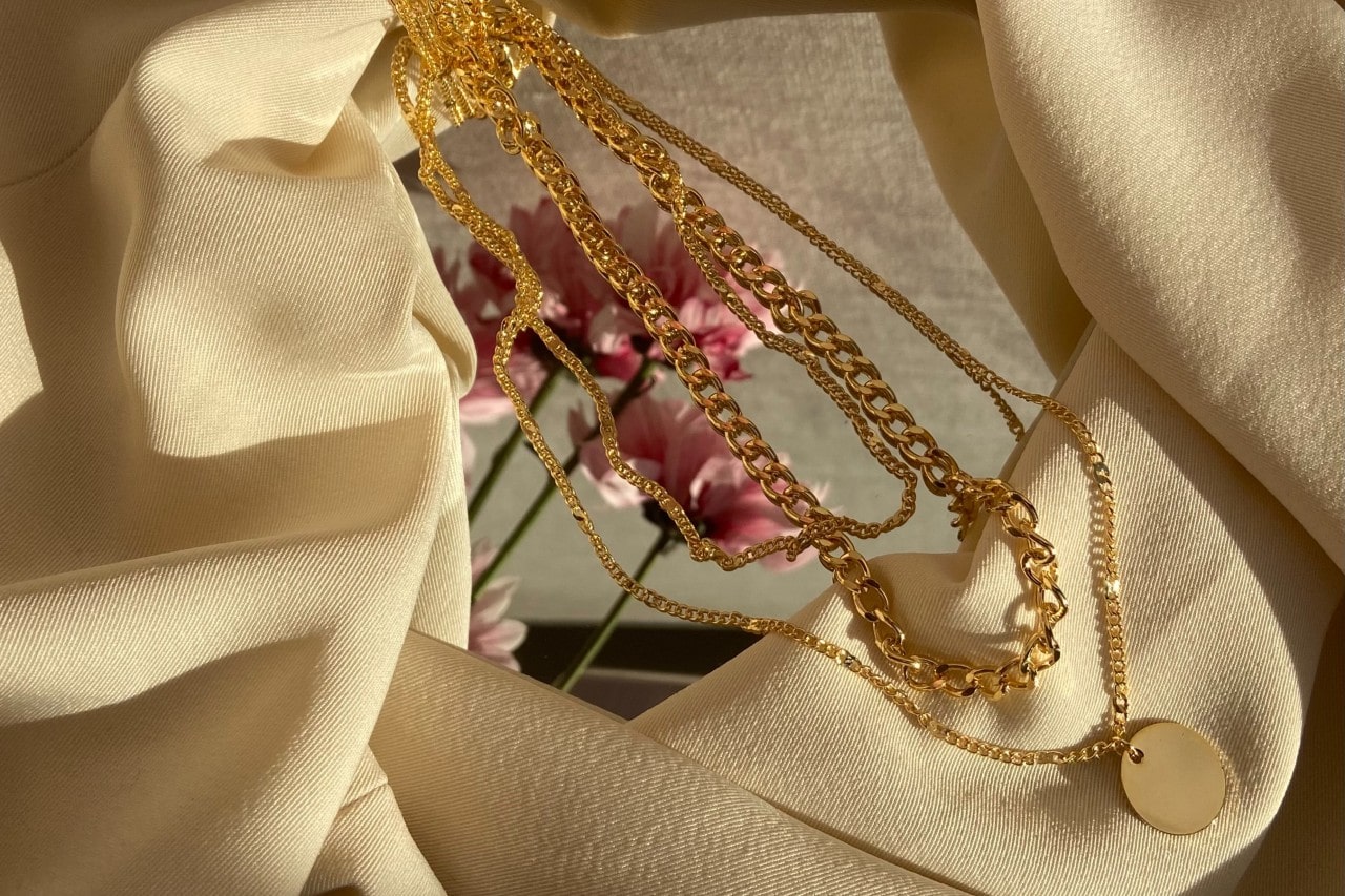 A set of layered gold necklaces displayed on cream fabric with a blurred pink flower in the background. A set of layered gold necklaces displayed on cream fabric with a blurred pink flower in the background.