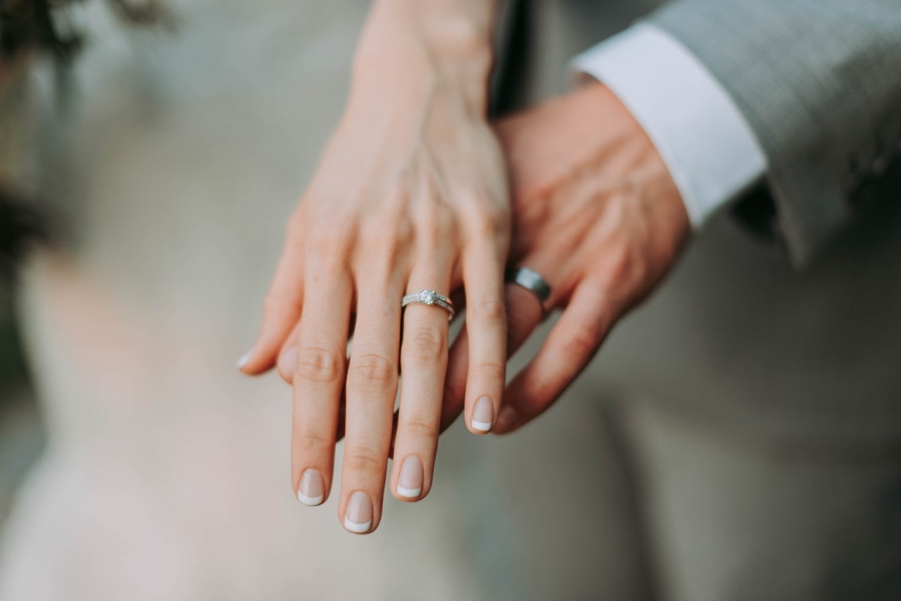 A close-up of two hands showing matching wedding bands, with the bride’s diamond ring and French-manicured nails in focus.