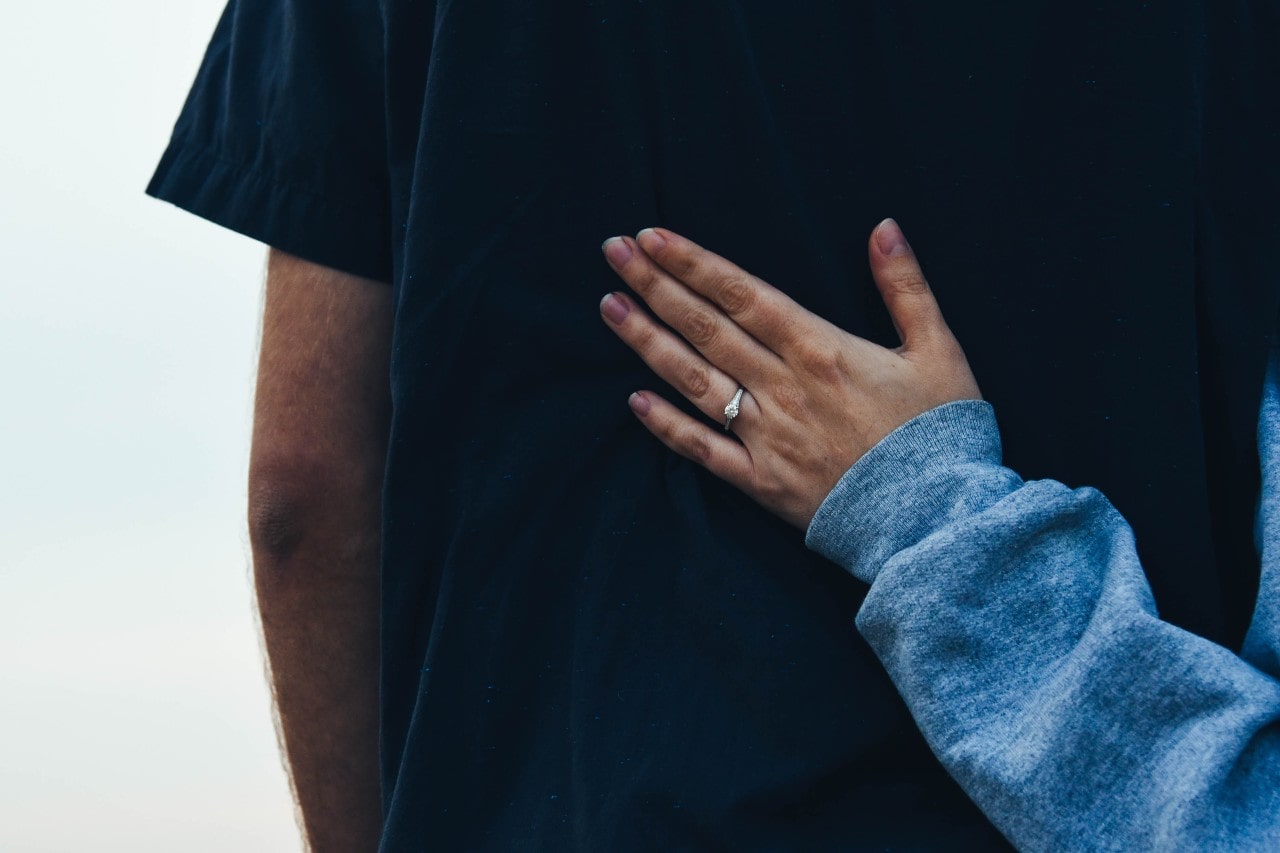 A woman wearing a blue sweatshirt and diamond engagement ring places her hand on a man’s back. A woman wearing a blue sweatshirt and diamond engagement ring places her hand on a man’s back.