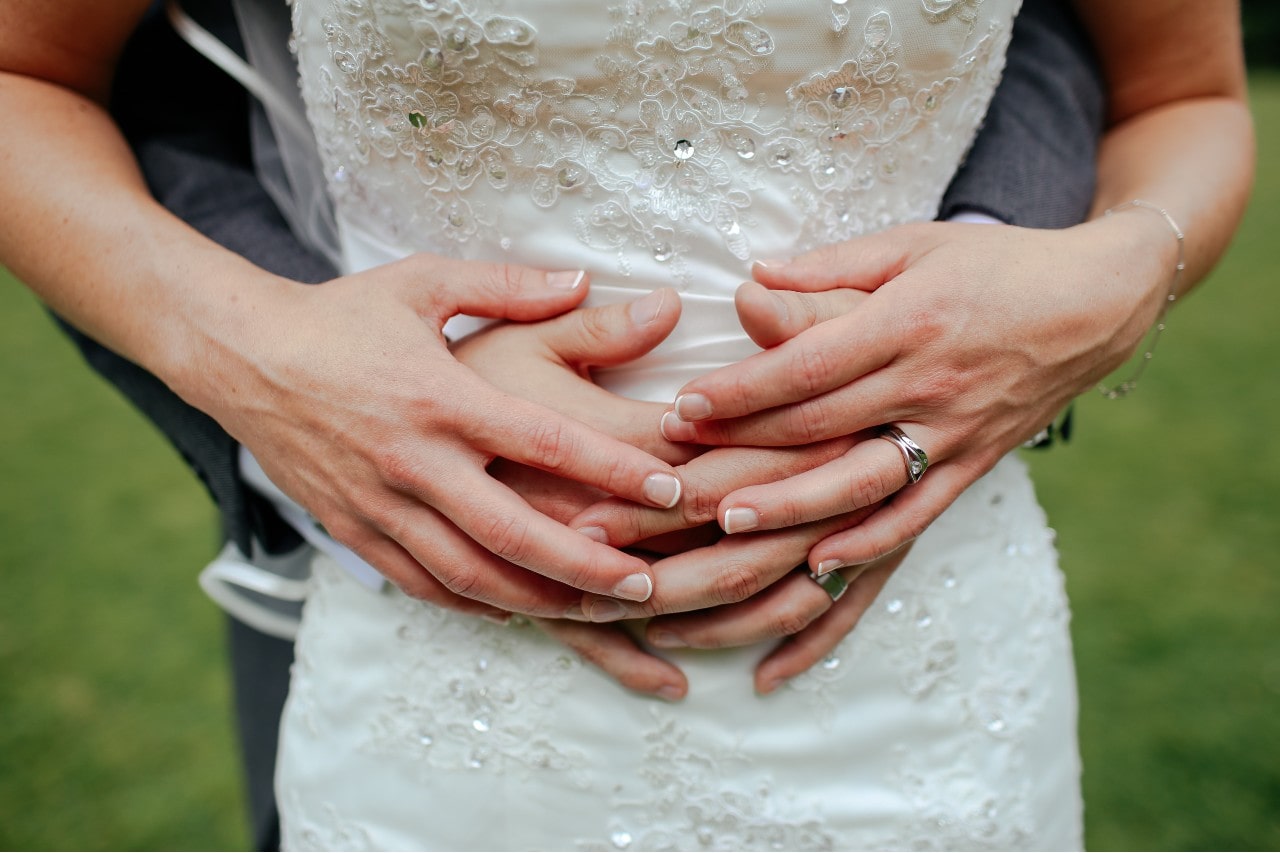 Close-up of a couple embracing, with hands gently resting on each other over a white, lace wedding dress. Close-up of a couple embracing, with hands gently resting on each other over a white, lace wedding dress.