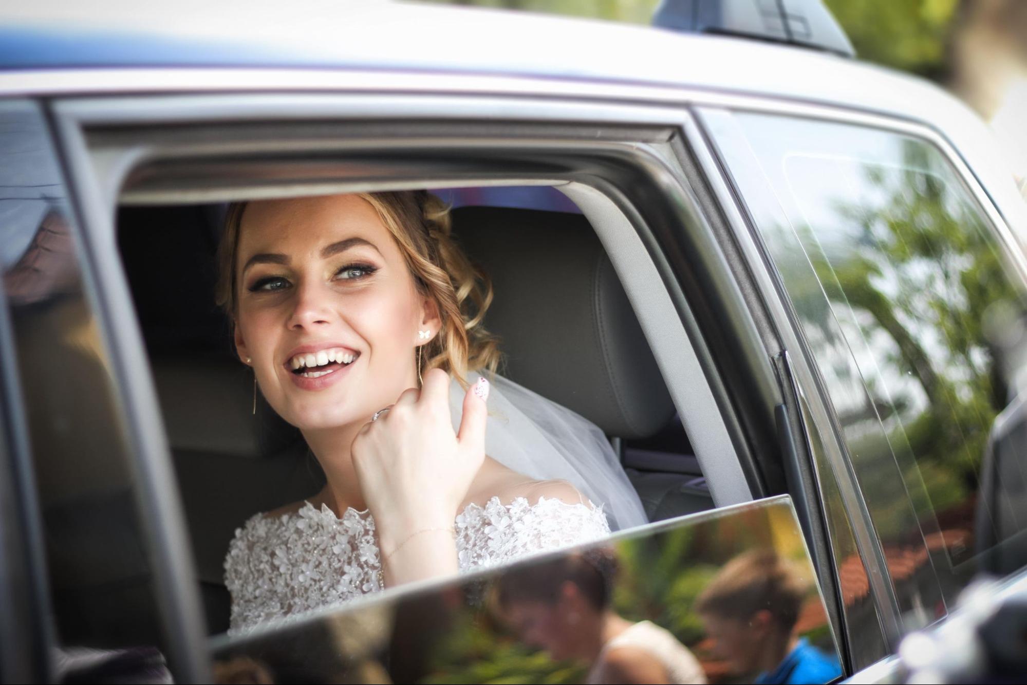 Smiling bride in a car, wearing a lace wedding dress, looking out the window with joy and excitement. Outside, blurred greenery and reflections are visible. Smiling bride in a car, wearing a lace wedding dress, looking out the window with joy and excitement. Outside, blurred greenery and reflections are visible.