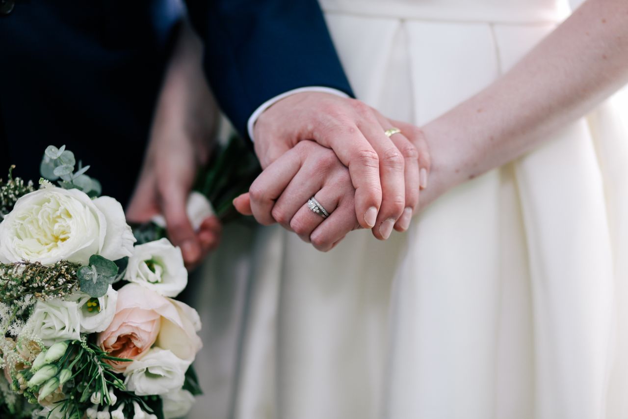 Close-up of a bride and groom holding hands, showing their wedding rings. The bride holds a bouquet of white and pink flowers, evoking a tender, romantic mood. Close-up of a bride and groom holding hands, showing their wedding rings. The bride holds a bouquet of white and pink flowers, evoking a tender, romantic mood.