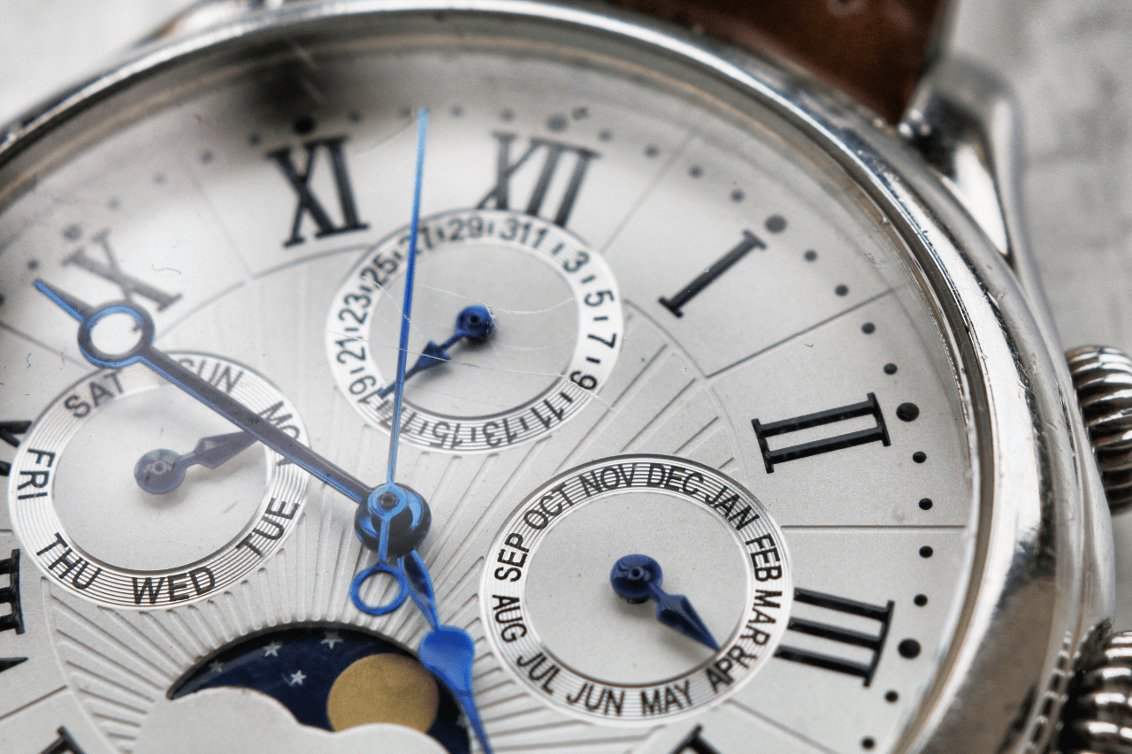 Close-up of a luxury wristwatch with Roman numerals, blue hands, and multiple subdials showing days, months, and moon phase. Close-up of a luxury wristwatch with Roman numerals, blue hands, and multiple subdials showing days, months, and moon phase.