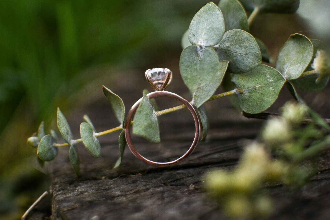 A rose gold solitaire engagement ring with a round diamond rests on a eucalyptus branch atop a rustic wooden surface.